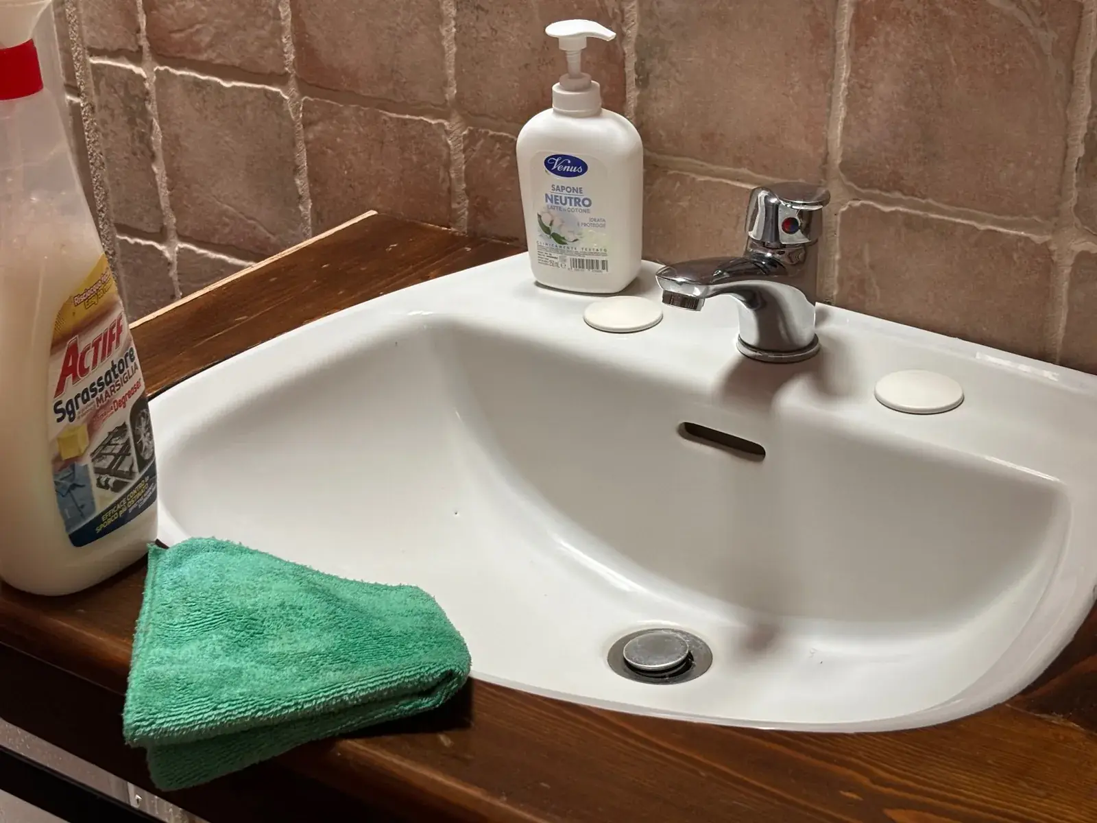 Lavabo in ceramica bianca con rubinetto cromato lucidato delle Antiche Dimore del Vescovado, accanto a sapone neutro e panno in microfibra sul piano in legno.